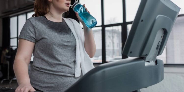 Mulher transpirando e se hidratando enquanto faz corrida em esteira ergométrica na academia