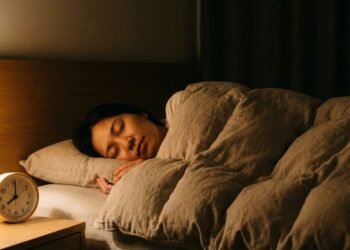 Fotografia realista de uma pessoa adulta dormindo tranquilamente em um quarto escuro e silencioso à noite. A iluminação é suave e quente, vinda de um abajur lateral. Em primeiro plano, sobre a mesa de cabeceira, um relógio visível marca 22h00. A cena transmite conforto, calma e sono profundo, sem a presença de eletrônicos