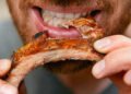 Close de um homem sorridente e com barba, segurando uma costela de porco inteira e grelhada com as duas mãos.
