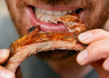 Close de um homem sorridente e com barba, segurando uma costela de porco inteira e grelhada com as duas mãos.