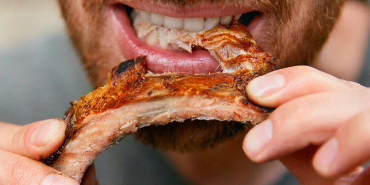 Close de um homem sorridente e com barba, segurando uma costela de porco inteira e grelhada com as duas mãos.