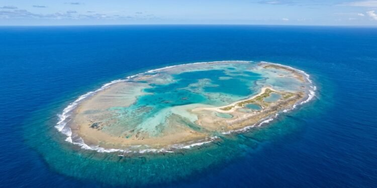 Vista aérea de um atol em formato circular, com recifes claros, lagoa de águas turquesa no centro e oceano azul-escuro ao redor.