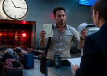 Passageiro segura passagem e documento diante de uma atendente no aeroporto, enquanto outros viajantes aguardam no chão com malas sob um grande relógio.