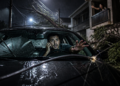 Homem dentro de um carro parado em rua alagada à noite segura um celular e estende a mão diante de um cabo de energia caído sobre o capô, sob chuva forte.