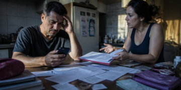 Homem olha preocupado para o celular enquanto uma mulher apresenta uma pasta com documentos, recibos e comprovantes espalhados sobre a mesa da cozinha.