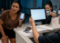 Mulher mostra no celular duas solicitações negadas enquanto atendentes apresentam uma maquininha e uma tela no balcão de uma clínica.