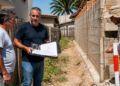 Três homens verificam a divisa entre terrenos enquanto um mede o muro com trena e outro mostra documentos da obra ao lado da construção.