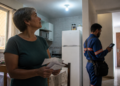 Mulher de cabelos grisalhos segura contas de energia na cozinha enquanto um técnico verifica o sistema elétrico da residência com um aparelho eletrônico.