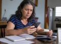 Mulher de 63 anos consulta o celular enquanto segura um comprovante e analisa anotações, documentos e cartões sobre a mesa de casa.