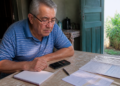 Homem idoso sentado à mesa observa um celular ao lado de caderno, caneta e documentos, em um ambiente residencial iluminado pela porta aberta.
