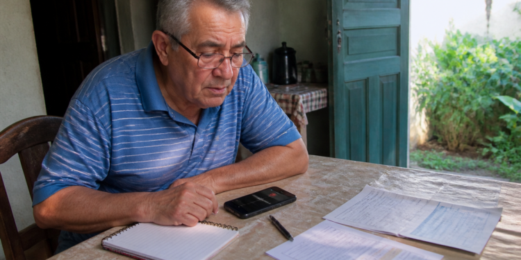 Homem idoso sentado à mesa observa um celular ao lado de caderno, caneta e documentos, em um ambiente residencial iluminado pela porta aberta.