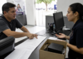 Cliente aponta para documentos de cobrança enquanto conversa com uma atendente diante de uma caixa com equipamento eletrônico.