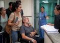 Família acompanha homem em cadeira de rodas enquanto mulher com celular conversa com atendente e profissional de saúde em corredor hospitalar.