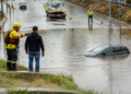 Bombeiro orienta um motorista diante de um carro parcialmente submerso em uma rua alagada, com agentes, cones e guincho ao fundo.