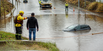 Bombeiro orienta um motorista diante de um carro parcialmente submerso em uma rua alagada, com agentes, cones e guincho ao fundo.