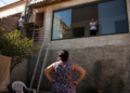 Mulher observa uma janela aberta para seu quintal enquanto dois trabalhadores realizam uma reforma na casa vizinha.