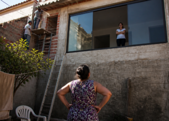 Mulher observa uma janela aberta para seu quintal enquanto dois trabalhadores realizam uma reforma na casa vizinha.