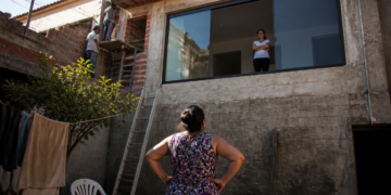 Mulher observa uma janela aberta para seu quintal enquanto dois trabalhadores realizam uma reforma na casa vizinha.