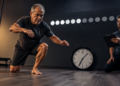 Homem mais velho realiza movimento de sentar e levantar do chão em uma academia, enquanto um avaliador observa com uma prancheta.