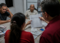 Família reunida à mesa analisa extratos, anotações e despesas médicas enquanto um homem idoso acompanha a conversa.