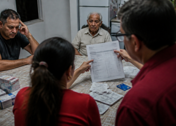 Família reunida à mesa analisa extratos, anotações e despesas médicas enquanto um homem idoso acompanha a conversa.