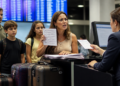 Mulher apresenta documentos a uma atendente no balcão de um aeroporto, acompanhada por um homem e duas crianças com malas, diante de painéis de voos.