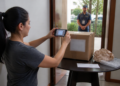 Mulher fotografa com o celular uma caixa de papelão sobre a mesa, enquanto um entregador aguarda do lado de fora da porta.