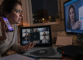 Mulher segura um celular e olha com preocupação para a tela de um monitor, ao lado de um notebook, roteador e anotações sobre a mesa em ambiente noturno.