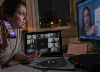 Mulher segura um celular e olha com preocupação para a tela de um monitor, ao lado de um notebook, roteador e anotações sobre a mesa em ambiente noturno.