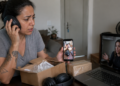 Mulher com expressão preocupada segura um celular enquanto usa fones de ouvido diante de uma caixa aberta, outro pacote e um notebook em videochamada.
