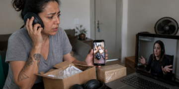 Mulher com expressão preocupada segura um celular enquanto usa fones de ouvido diante de uma caixa aberta, outro pacote e um notebook em videochamada.