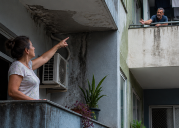 Mulher aponta para infiltração e manchas de umidade na sacada enquanto discute com um vizinho no apartamento acima.
