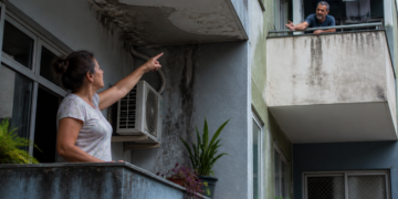 Mulher aponta para infiltração e manchas de umidade na sacada enquanto discute com um vizinho no apartamento acima.