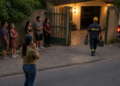 Técnico com capacete caminha em direção a uma casa enquanto moradores aguardam no portão e uma mulher fala ao telefone na calçada, ao lado de uma van de serviço.