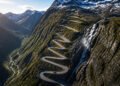Vista aérea da estrada Trollstigen subindo a montanha em curvas fechadas, ao lado de uma cachoeira e cercada por picos com neve.