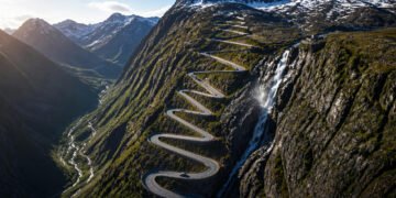 Vista aérea da estrada Trollstigen subindo a montanha em curvas fechadas, ao lado de uma cachoeira e cercada por picos com neve.