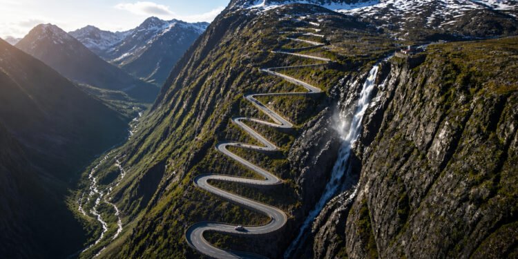 Vista aérea da estrada Trollstigen subindo a montanha em curvas fechadas, ao lado de uma cachoeira e cercada por picos com neve.