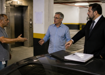 Três homens discutem ao lado de um carro em uma garagem de condomínio, com documentos sobre o veículo e vagas ao fundo.