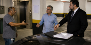 Três homens discutem ao lado de um carro em uma garagem de condomínio, com documentos sobre o veículo e vagas ao fundo.
