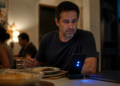 Homem sentado à mesa durante o jantar observa um celular iluminado, com prato de comida e notebook fechado ao lado.