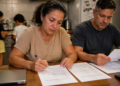 Casal analisa contratos e cobranças sobre uma mesa, com notebook e calculadora, enquanto duas crianças permanecem na cozinha ao fundo.
