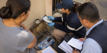 Técnica com luvas registra dados em um tablet ao lado de um hidrômetro, enquanto uma moradora e outro profissional acompanham a inspeção com documentos.