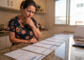 Mulher em uma cozinha observa várias contas alinhadas sobre a bancada, com expressão de preocupação, ao lado de uma calculadora e um suporte de mesa.