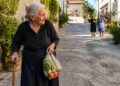Mulher idosa com bengala e sacola de compras caminha por uma rua de vila, enquanto duas pessoas conversam ao fundo.