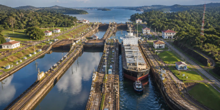 Vista aérea das eclusas do Canal do Panamá com um grande navio cargueiro sendo conduzido entre câmaras de concreto cercadas por vegetação.
