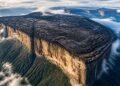 Vista aérea de uma montanha de topo plano com paredões verticais, cercada por nuvens e floresta, com cachoeiras escorrendo pelas bordas.