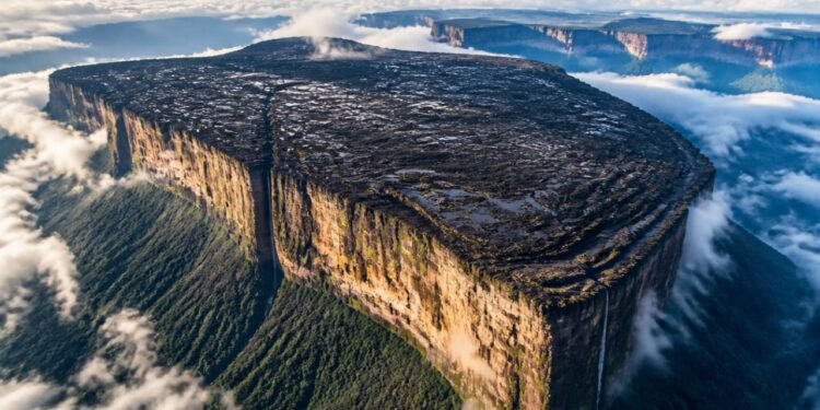 Vista aérea de uma montanha de topo plano com paredões verticais, cercada por nuvens e floresta, com cachoeiras escorrendo pelas bordas.
