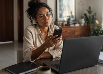 Mulher sentada à mesa observa o celular diante de um notebook aberto, com papéis, caderno e xícara ao redor, em um ambiente doméstico de trabalho.