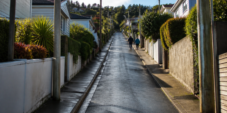 Rua residencial estreita e muito íngreme sobe entre casas, muros, jardins e postes, enquanto duas pessoas caminham pela calçada.