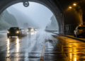 Veículos circulam por uma pista molhada na saída de um túnel da Rodovia dos Imigrantes, com neblina, vegetação e carro de serviço no acostamento.